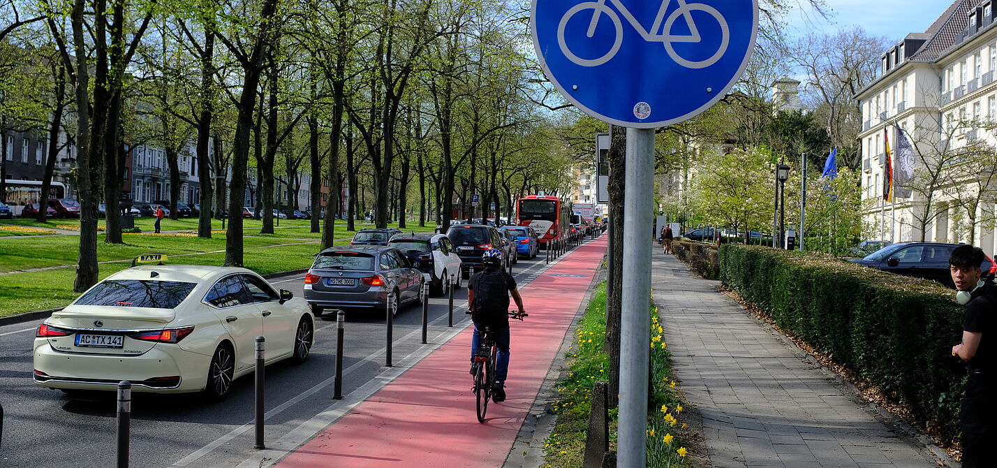 Landesversammlung_ADFC_NRW_2025._ADFC_NRW__B._Glitz___96_.JPG Ein geschützter Radfahrstreifen mit Roteinfärbung. Radwegschild und einer Radfahrerin.