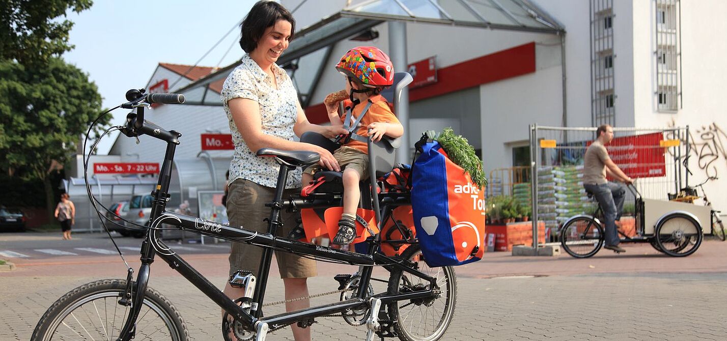 Kindertransport im Kindersitz hinten Frau schnallt Kind im Kindersitz an. Es sitzt hinten auf dem Fahrrad, dahinter sind noch Gepäcktaschen angehängt.