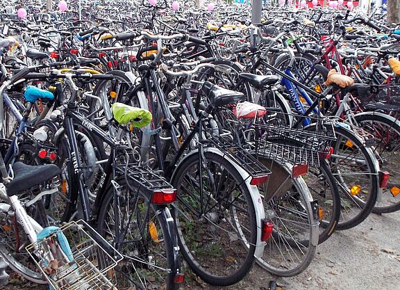 Fahrradparkplatz ohne Abstellanlagen Hunderte von Fahrrädern verschiedener Farben und Modelle stehen dicht gedrängt und ungeordnet auf einer befestigten Fläche; die Räder sind ineinander verschachtelt, mit Körben, Taschen und bunten Sattelbezügen ausgestattet, ohne erkennbare Parkordnung oder Abstellvorrichtungen.