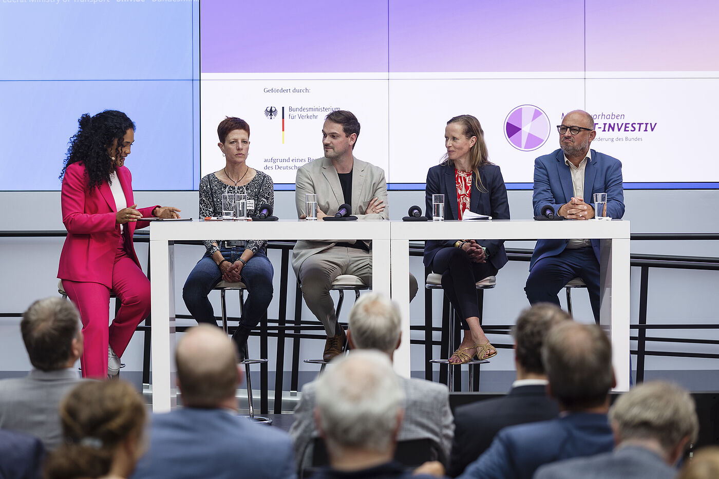 Podiumsdiskussion Podiumsdiskussion mit fünf Personen an weißen Tischen vor lila Hintergrund, Moderatorin in pinkem Anzug links, vier Diskussionsteilnehmende rechts, darunter Mann in beigem Anzug und Mann in blauem Anzug ganz rechts, Publikum sitzt davor