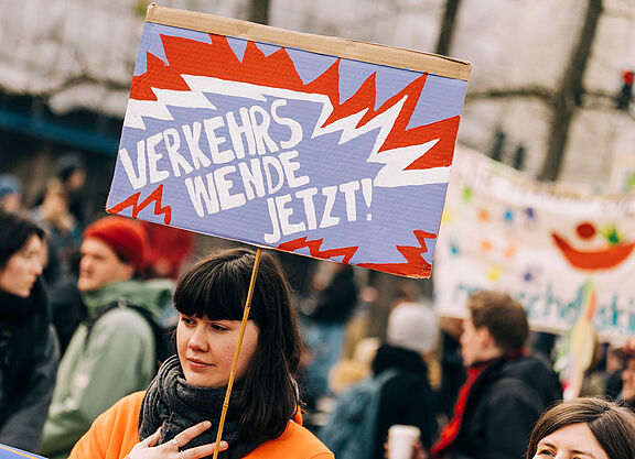 Dossier Junge Menschen Verkehrswende Jetzt! Junge Frau auf einer Demo hält ein Schild in die Luft, auf dem steht "Verkehrswende Jetzt!"
