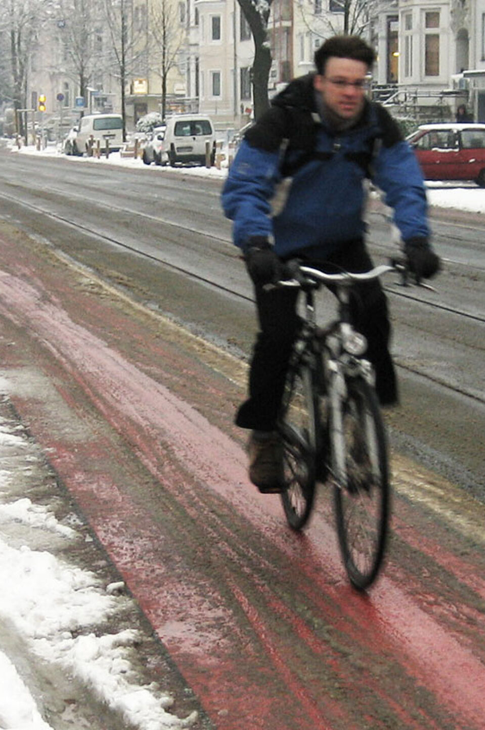 Radfahren im Winter Ein Radfahrer in Winterkleidung fährt auf einem rot markierten Radfahrstreifen neben einer Straße, der weitgehend von Schnee befreit ist, während am Straßenrand und auf geparkten Autos Schnee liegt.